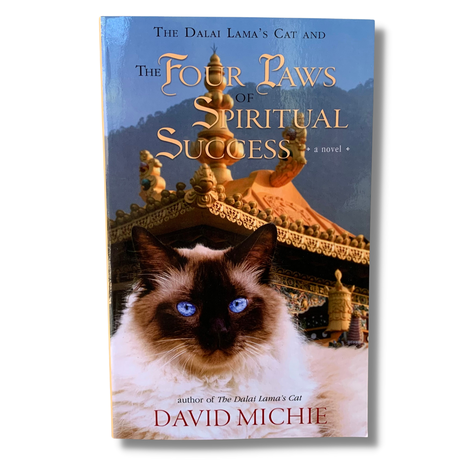 A book cover featuring a cat with blue eyes in the foreground with a blurred image of a person in a traditional robe in the background. The title of the book is visible and reads: 'The Dalai Lama's and the Four Paws of Spiritual Success - A Novel'.