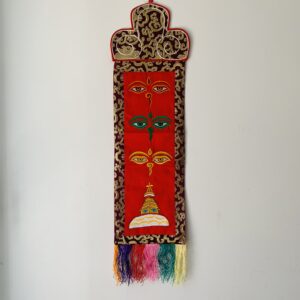 A red brocade wall hanging featuring embroidered Buddha's Wisdom eyes and a Kathmandu Boudhanath Stupa design, with tassels at the bottom.