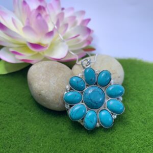 A howlite turquoise flower pendant with white metal setting, placed on a green leaf-like surface next to a rock.