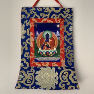 A Mini Brocade Thangka depicting the Medicine Buddha with a red and gold frame, hanging against a blue background.