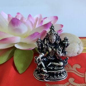 A silver-plated Green Tara statue seated on a red surface with a large pink flower behind it and a small rock in front.