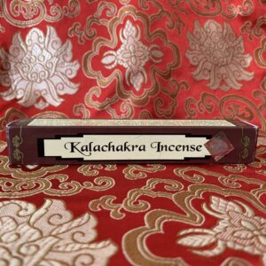 A box of Kalachakra incense placed on a red patterned surface. The box is brown with a small window displaying the product name.