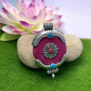 A round dark purple amulet pendant with turquoise and red coral like pieces, hand-decorated and set in a silver-colored frame on a green background.