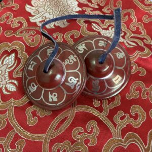 A pair of red brass meditation cymbals with the Mani Mantra written in white. They are placed on a red and gold patterned surface.