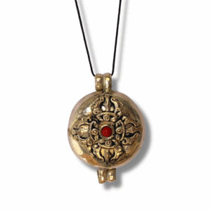 Round Tibetan-style brass gau pendant with raised double vajra (dorje) motif and red stone centre, suspended on a black cord necklace.